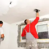 Dwóch mężczyzn w ogrodniczkach tynkuje sufit i ściany w pomieszczeniu.