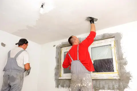 Dwóch mężczyzn w ogrodniczkach tynkuje sufit i ściany w pomieszczeniu.